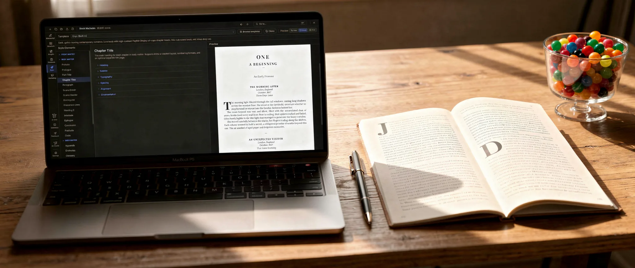 A laptop running a manuscript formatting app beside an open printed book showing chapter-opener drop caps, with a pen and a jar of colorful candy on a warm wooden desk.